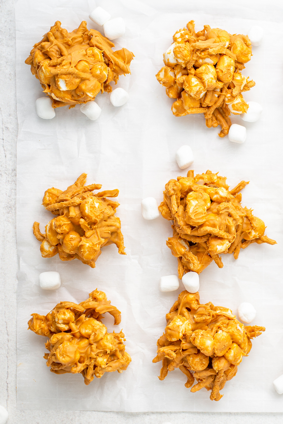 Six haystack cookies on a sheet of wax paper. Mini marshmallows are scattered around them.