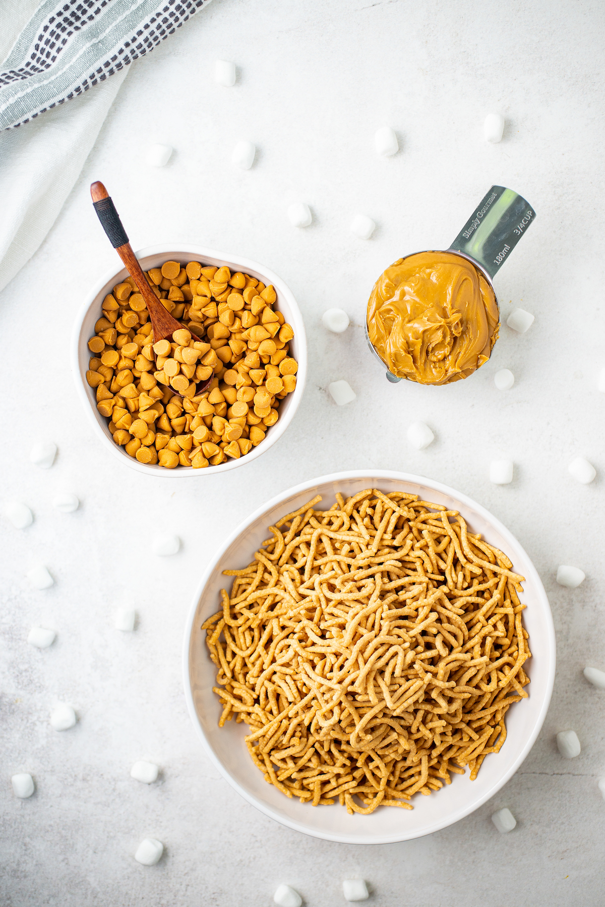 Dishes of butterscotch chips, peanut butter, and crispy chow mein noodles on a cookie sheet lined with wax paper. Mini marshmallows are dotted around the dishes.
