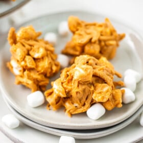 Three no-bake peanut butter haystacks on a plate, with marshmallows scattered over them.