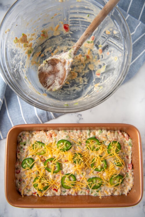 An empty mixing bowl with a wooden spoon inside, next to a loaf pan with unbaked bread inside. Sliced jalapenos and shredded cheddar have been sprinkled on top of the dough.