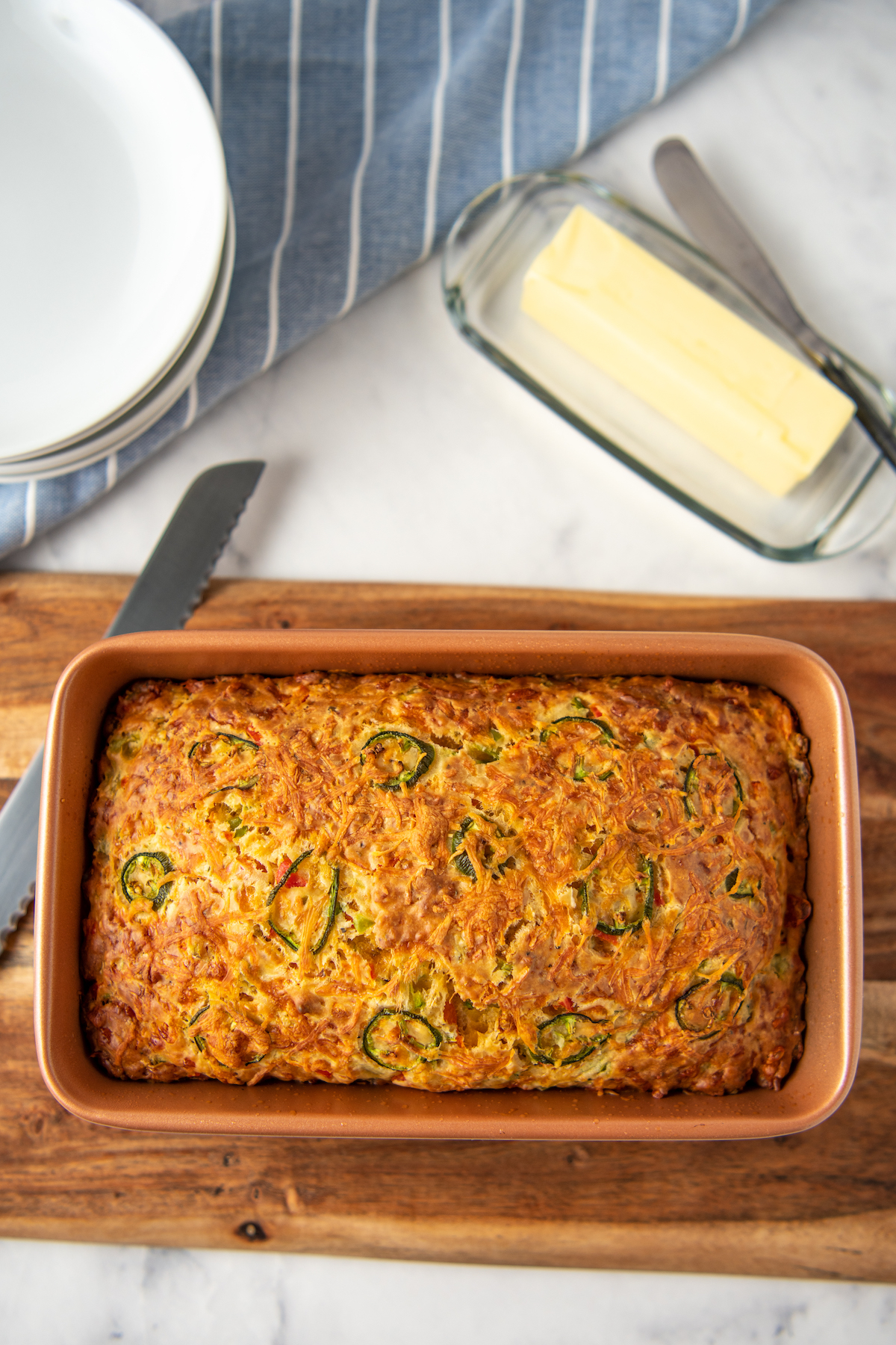 Baked jalapeno cheddar bread in a loaf pan, next to a dish of butter and a bread knife.