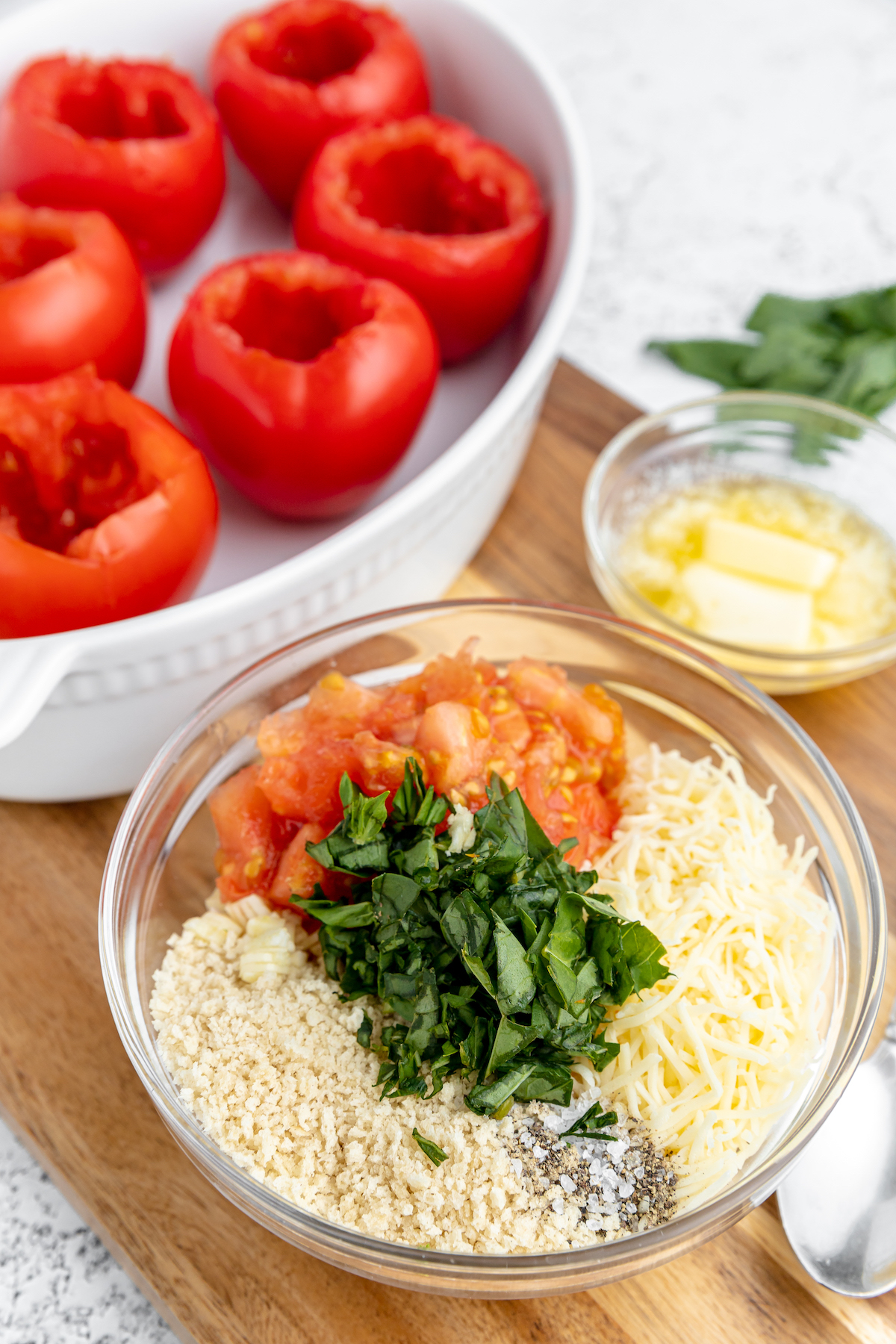 A white oval baking dish with cored tomatoes inside. A small glass bowl filling ingredients is nearby on the table.