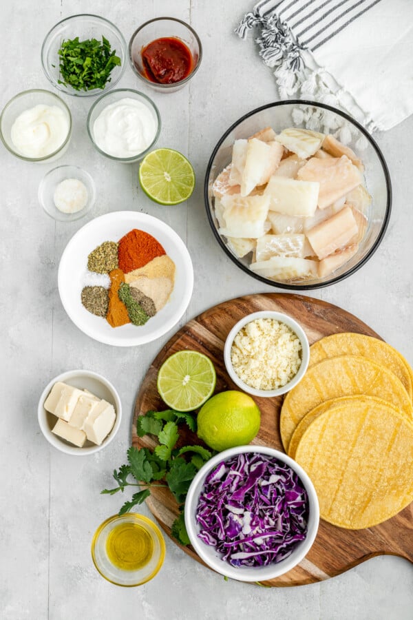 Sour cream, mayonnaise, lime juice, half a lime, raw fish fillets, spices and seasonings, butter, cilantro, cotija cheese, purple cabbage shreds and corn tortillas laid out on the counter.