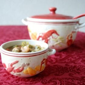 Small white bowl filled with shrimp gumbo in front of white bowl with red cap and red spoon ladle on a red patterned table cloth
