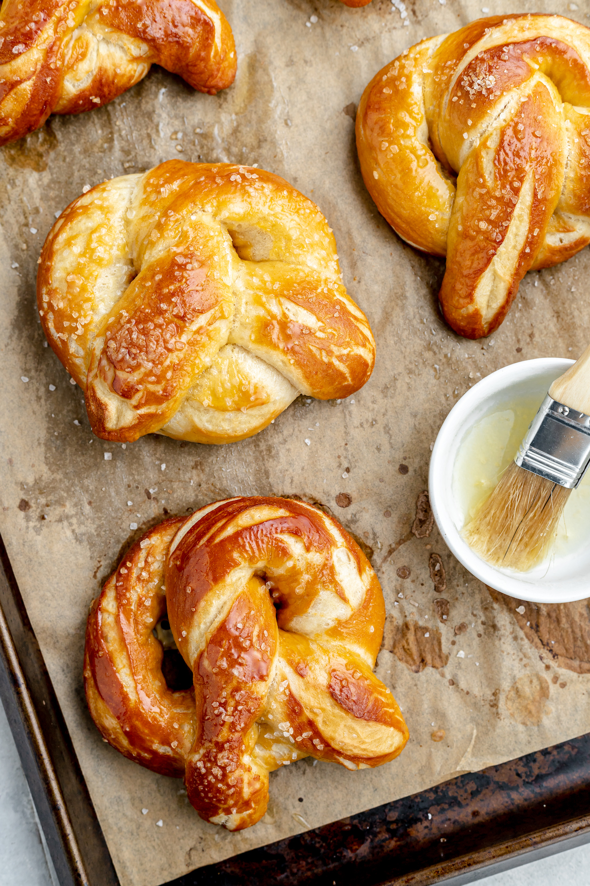 Baked pretzels on a parchment-lined baking sheet with a dish of egg wash and a brush.