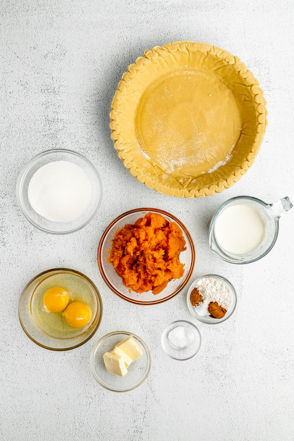 An unbaked pie crust, buttermilk, sweet potato puree, sugar, eggs, butter, salt, cinnamon, nutmeg and baking soda laid out on the counter.