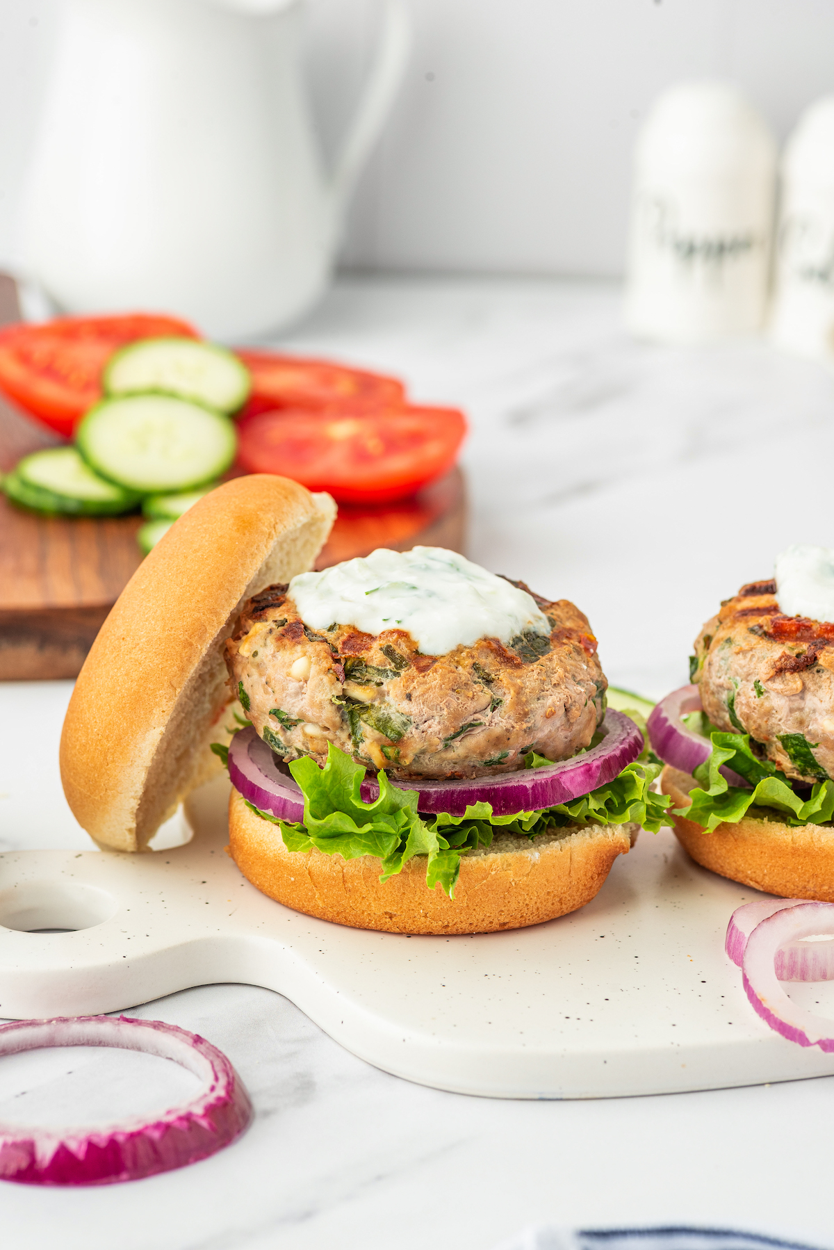 A Greek turkey burger, with the top bun removed, topped with creamy sauce. Another burger is nearby on a cutting board, while a wooden tray of sliced tomatoes and onions is in the background of the shot.