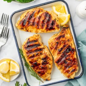 An enamel tray with grilled chicken breast, surrounded by ingredients like broccoli, garlic, lemon wedges, olive oil, and rosemary.