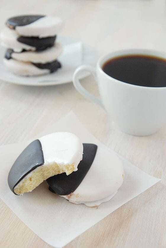 Two black and white cookies stacked on a square of parchment paper next to a cup of coffee