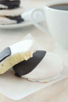A black and white cookie with a bite taken out of it stacked on top of a whole cookie with a cup of coffee and a second stack of cookies in the background