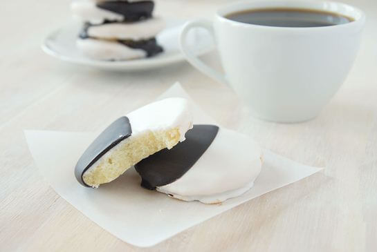 A black and white cookie with a bite taken out of it stacked on top of a whole cookie with a cup of coffee and a second stack of cookies in the background