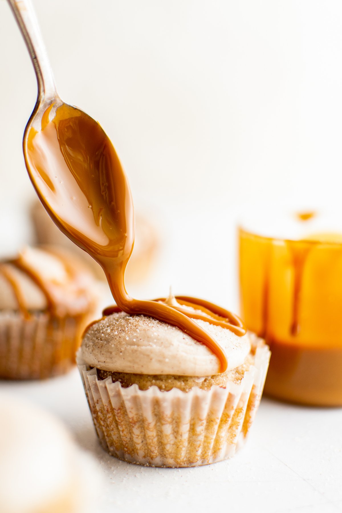A churro cupcake next to a container of caramel sauce, with sauce being drizzled over the cupcake with a teaspoon.