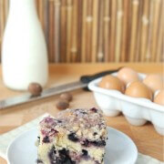 Blueberry buckle on a white plate with eggs, milk and nutmeg in background.