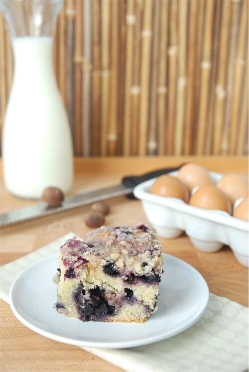 Blueberry buckle on a white plate with eggs, milk and nutmeg in background.