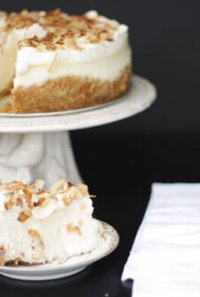 Slice of coconut cheesecake on a plate in front of whole cheesecake on a cake stand with a white napkin and fork