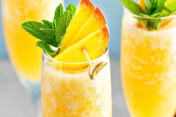Up close photo of peach bellini in a glass with fresh peaches and mint leaves.