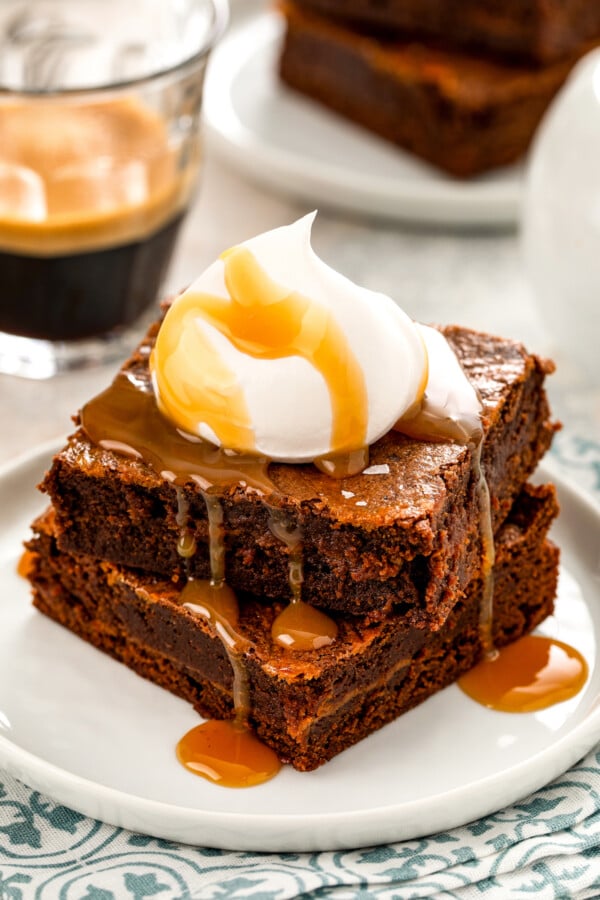 Two rich and fudgy salted caramel brownies stacked on top of each other with a dollop of whipped cream on top and a drizzle of more caramel sauce.