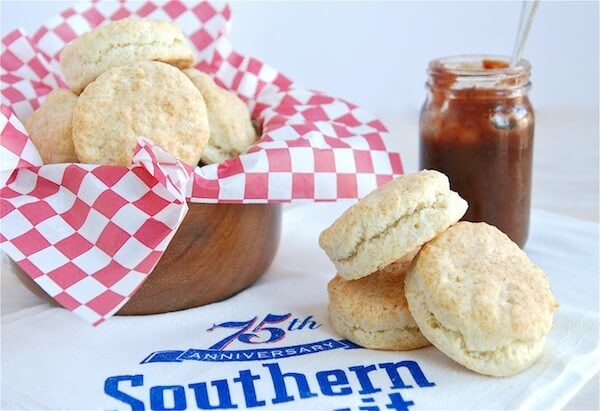 Three buttermilk biscuits on a napkin with some more placed in a bowl with a jar of jam with a spoon inside.