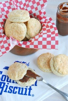 Buttermilk biscuits in a bowl with some on a towel sliced in half with jam inside.