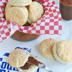 Buttermilk biscuits in a bowl with some on a towel sliced in half with jam inside.