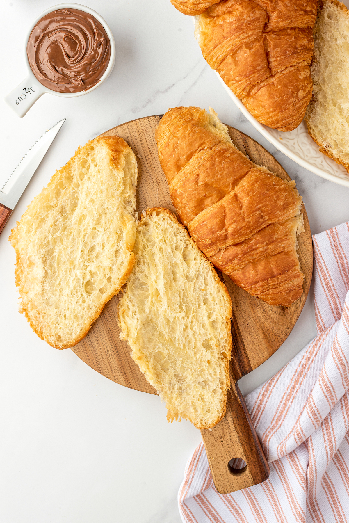 A cutting board with one whole croissant, and one halved croissant. More croissants and a cup of Nutella are nearby.