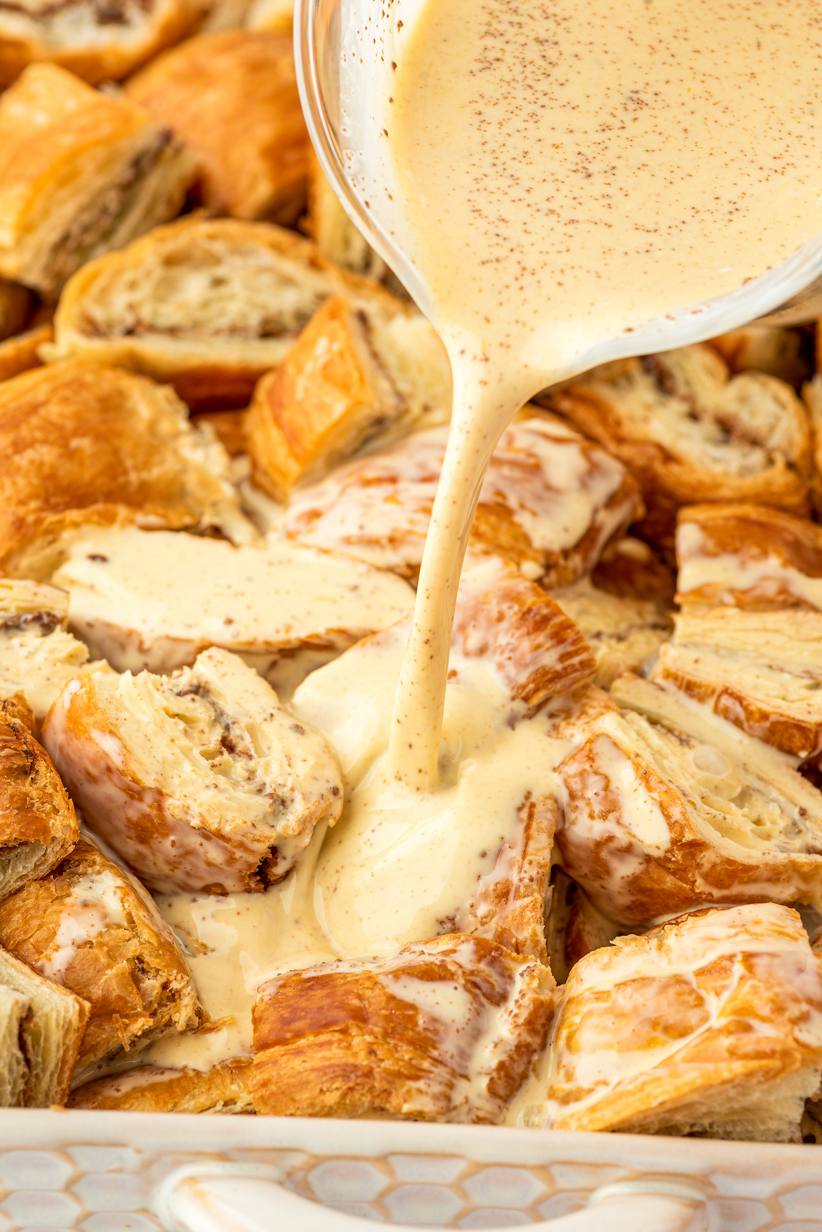 Homemade custard mixture being poured over croissants in a baking dish.