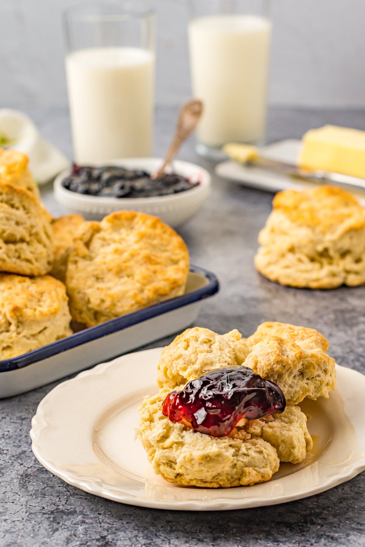 A biscuit split on a small plate, and topped with jelly. In the background of the shot are glasses of milk, a bowl of jelly, a butter dish, and a tray of additional biscuits.