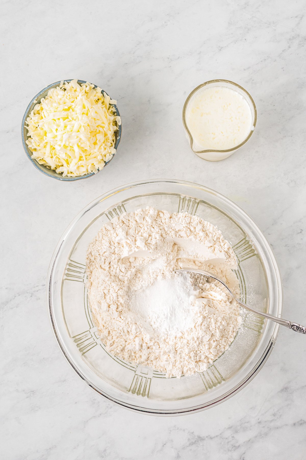 A large glass mixing bowl with dry baking ingredients, next to a smaller bowl of grated butter and shortening, and a measuring cup of buttermilk.