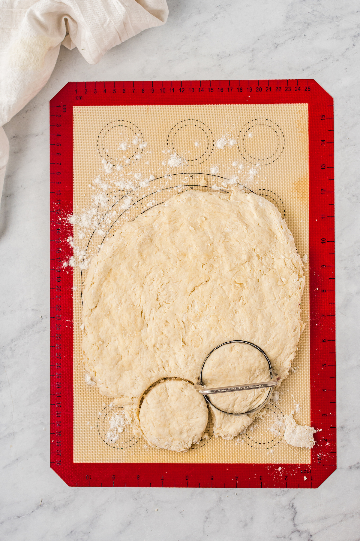 A circle of biscuit dough on a baking mat. One biscuit has been cut from the dough, and the biscuit cutter is resting on the remaining dough.