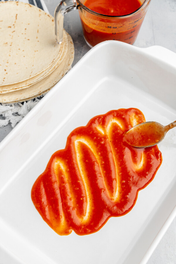 Red sauce being spread in the bottom of a rectangular white baking dish.