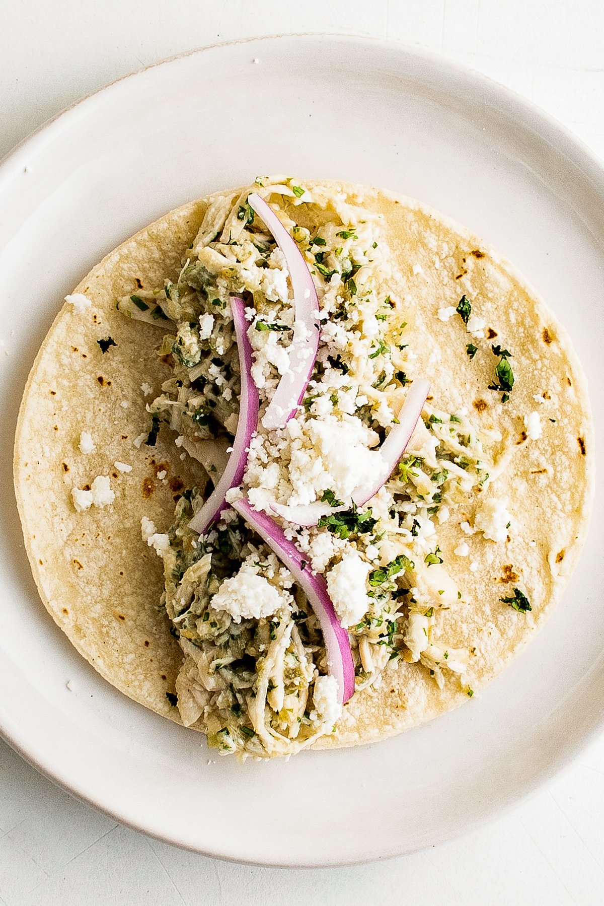 A flour tortilla topped with shredded salsa verde chicken, red onion, and cotija cheese.