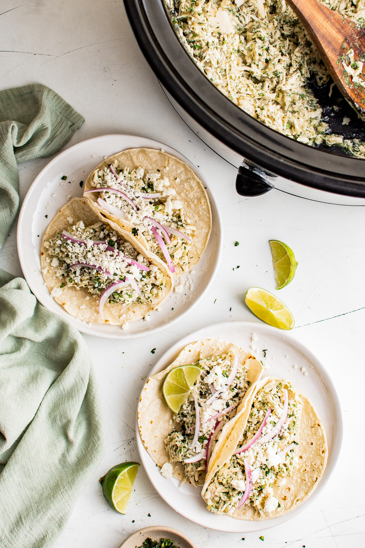 Plated salsa verde chicken tacos with toppings, next to a slow cooker insert with more salsa verde chicken inside.