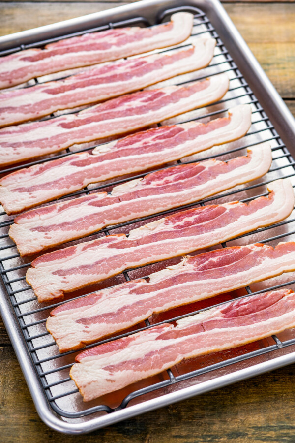 Showing how to cook bacon in the oven by lining strips of uncooked bacon lined on top of a wire rack over a baking sheet.