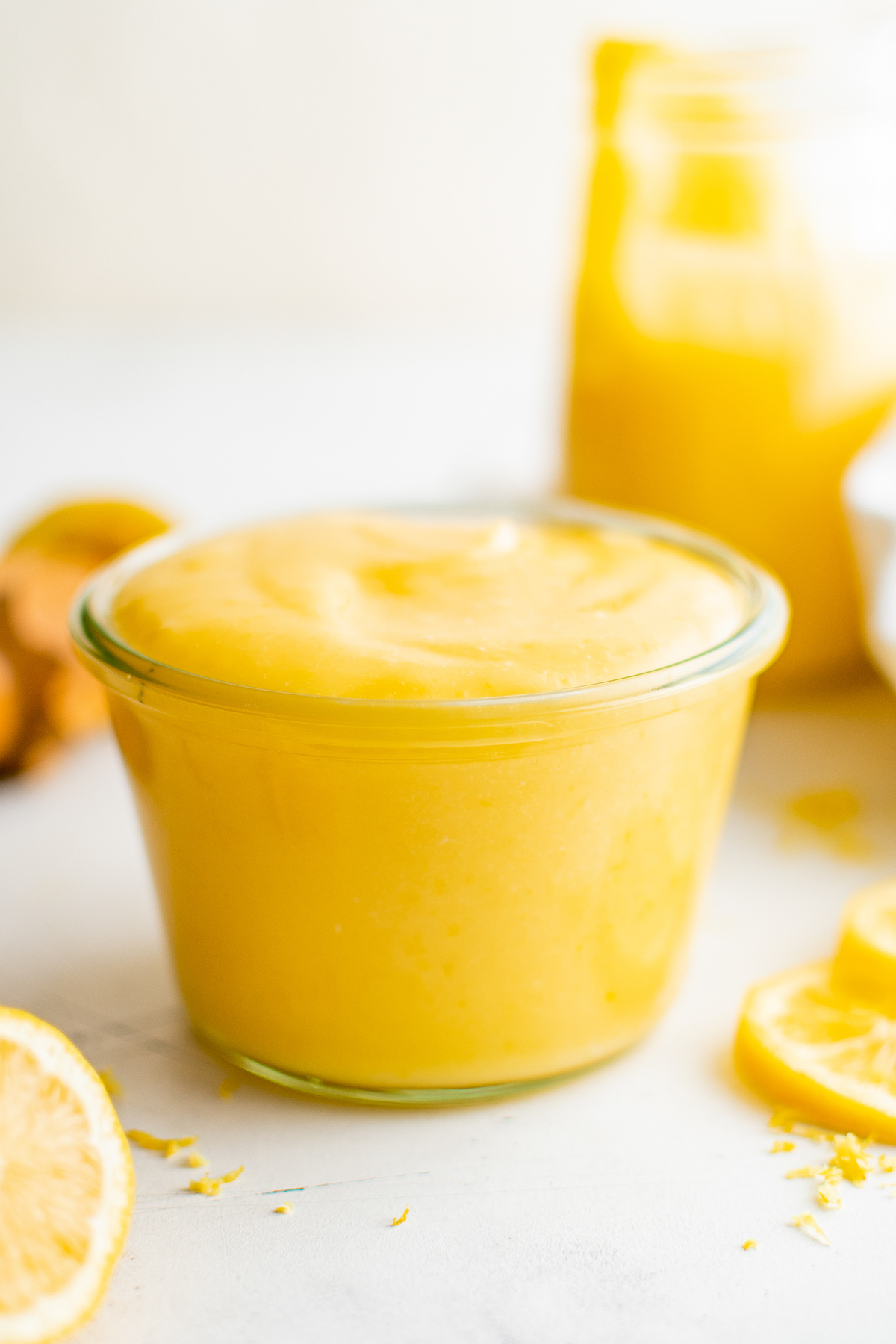 A small glass cup of creamy lemon curd. Lemon slices and a larger jar of lemon curd are in the background.