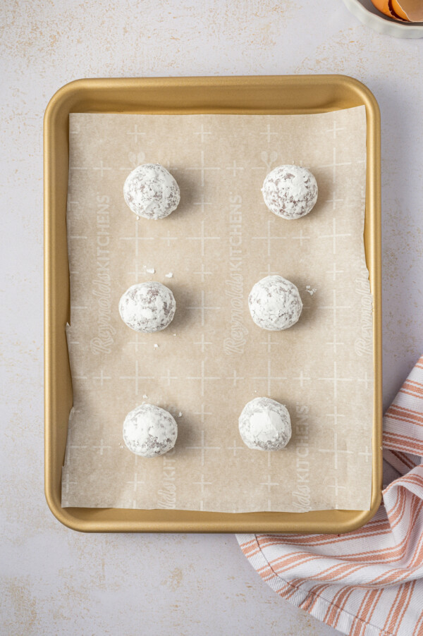 Balls of cookie dough coated in powdered sugar, lined up on a baking sheet.