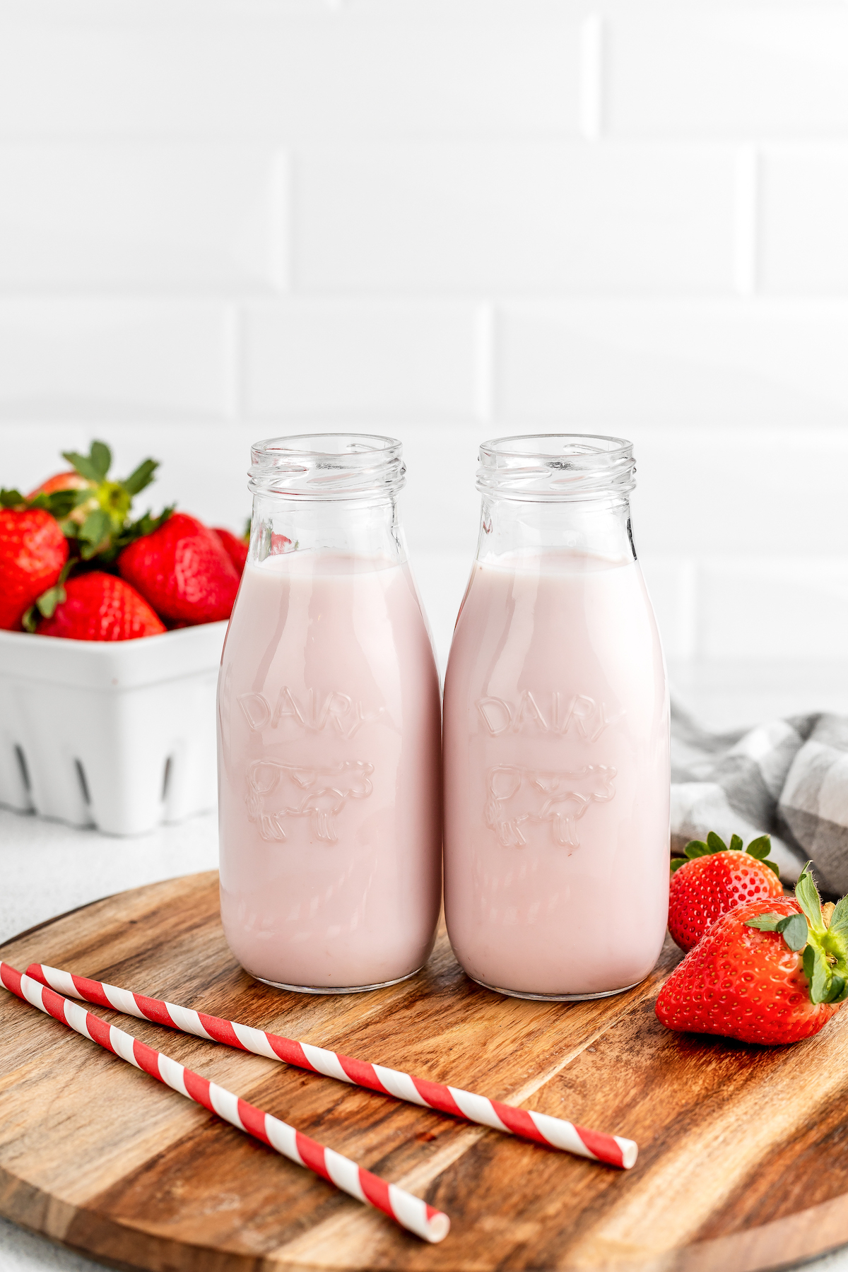 Pale pink milk in two small glass milk jars. Strawberries and paper straws are on the table as well.