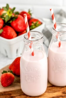 Two servings of strawberry milk, with red-and-white straws. A pint of fresh strawberries is in the background of the shot.