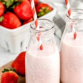 Two servings of strawberry milk, with red-and-white straws. A pint of fresh strawberries is in the background of the shot.