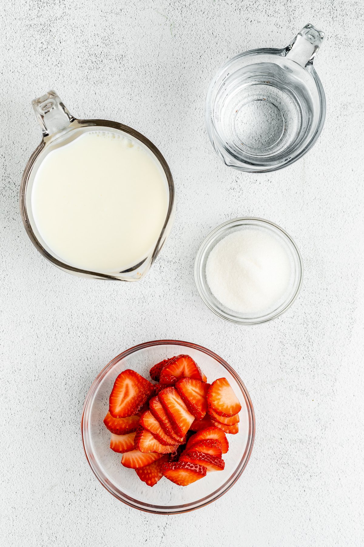 Milk, water, sugar, and sliced strawberries, on a white work surface.