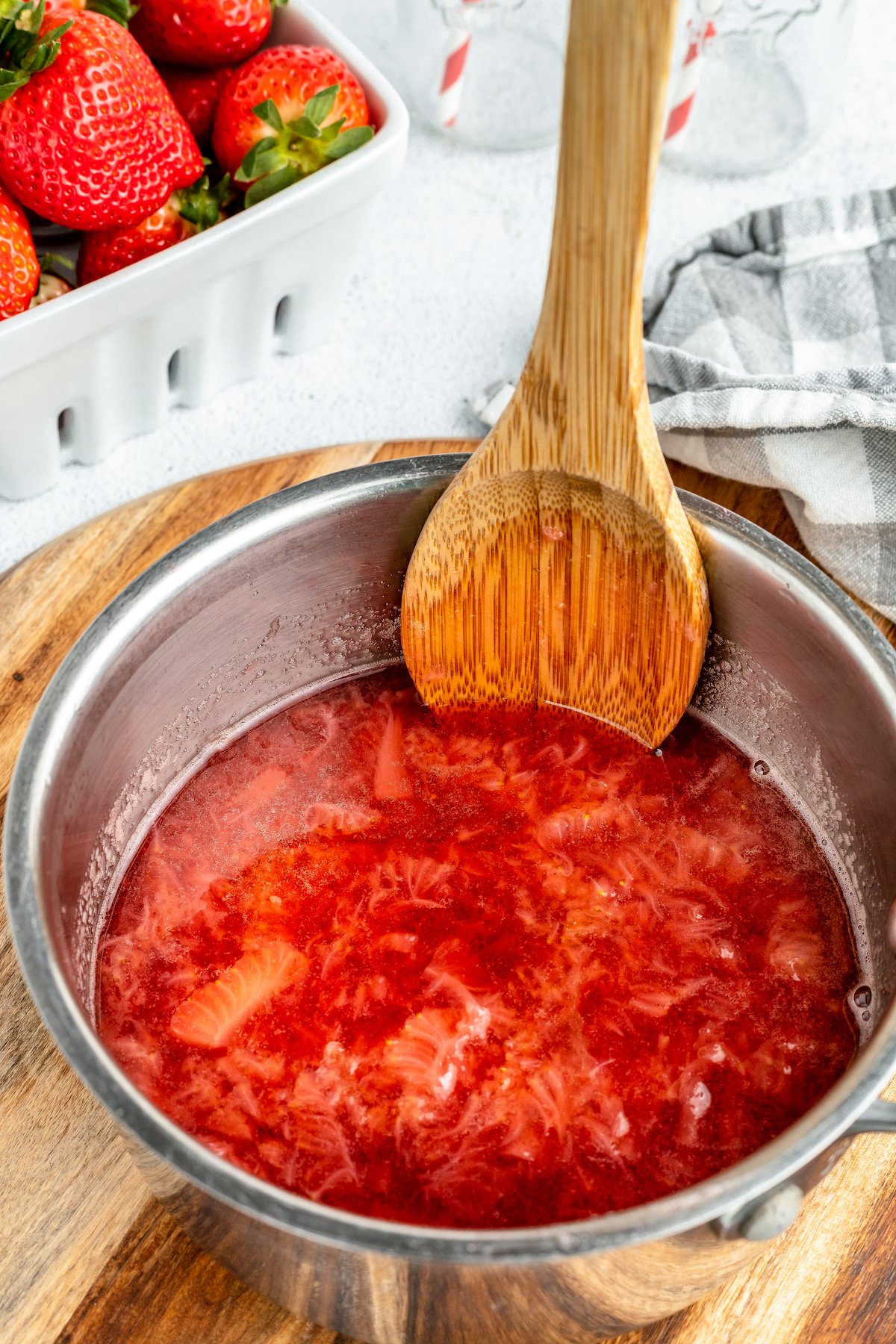 Simmering strawberries in a small saucepan. A wooden spoon is resting in the saucepan as well.