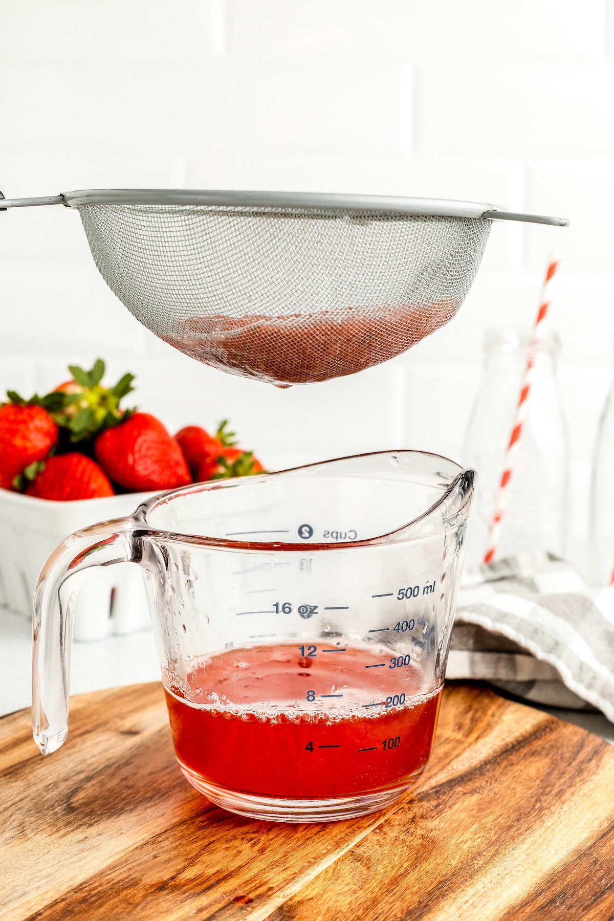 A strainer is held over a measuring cup full of red syrup.