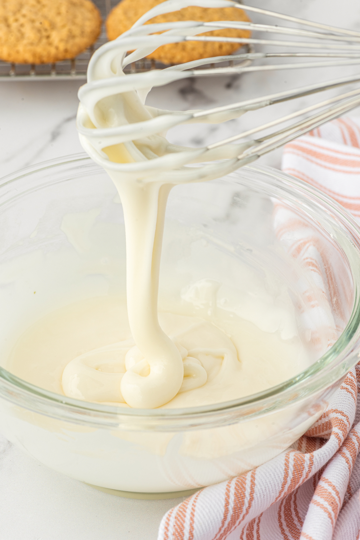 A small glass bowl of thick white icing, with a whisk drizzling some of the icing into the bowl.