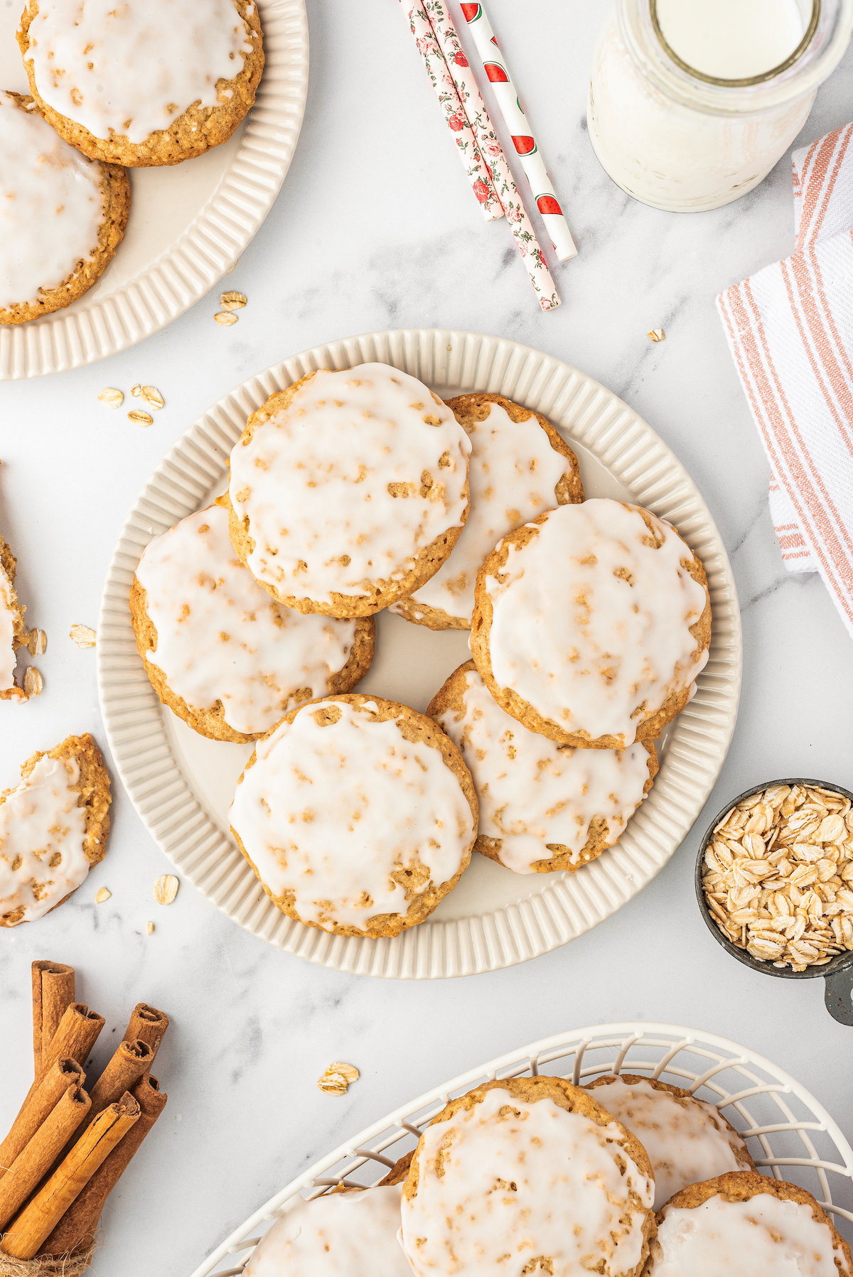 Several plates of cookies on a table, with a glass of milk, paper straws, cinnamon sticks, and cloth napkins.