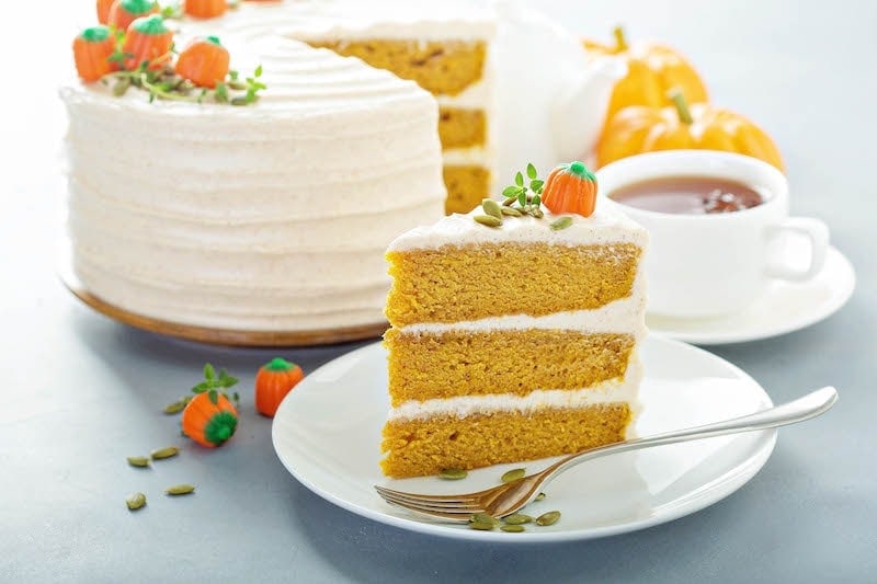 A slice of pumpkin cake in front of a frosted layer pumpkin cake with a slice taken out.