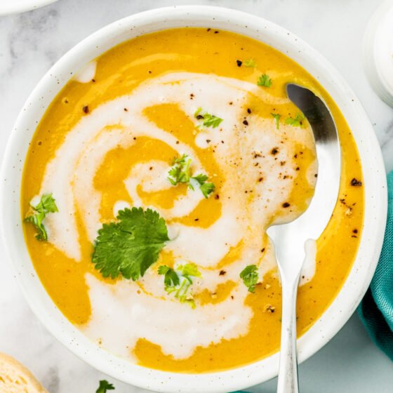 a bowl of butternut squash coconut curry soup with a cream swirl and herb garnish with a spoon in it