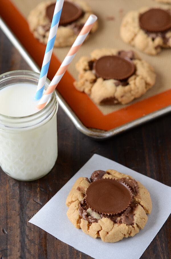 A mason jar filled with milk sits next to a cookie on a white square paper that's topped with a large peanut butter cup 