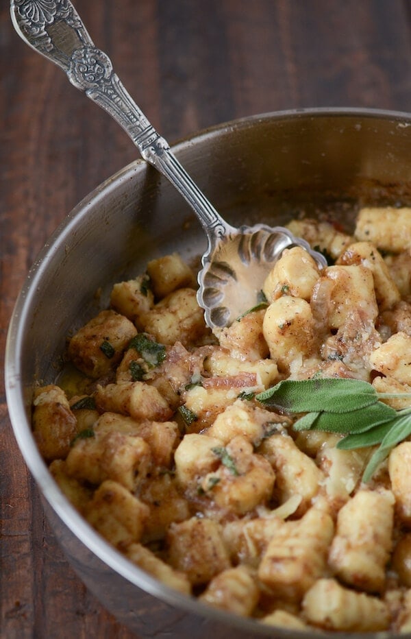 A Bowl of Potato Gnocchi with Brown Butter Sage Sauce on a Wooden Table with a Metal Spoon Digging Into the Gnocchi