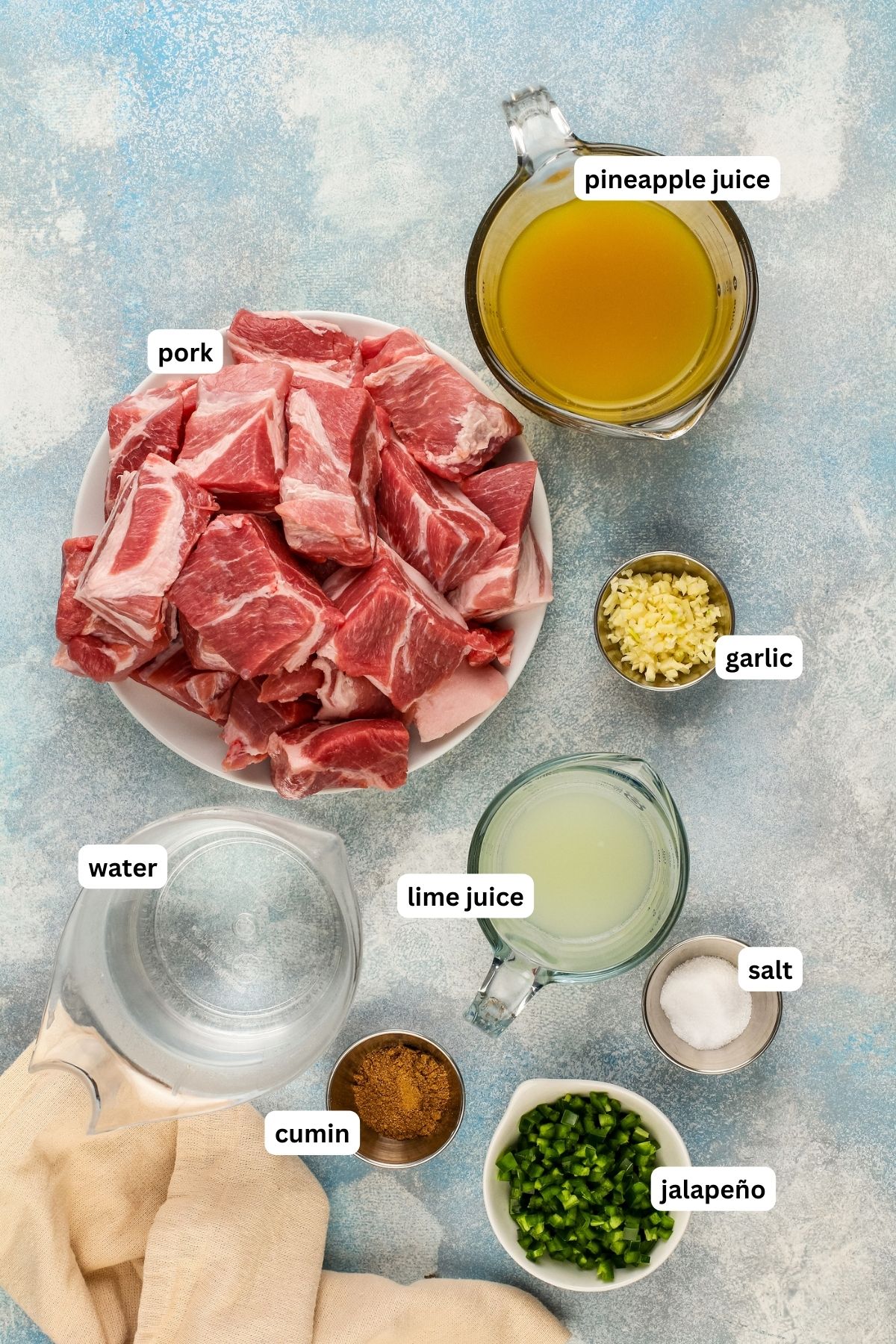 Labeled ingredients for pork carnitas recipe arranged in bowls. From top to bottom: pineapple juice, cubed pork, fresh garlic, water, lime juice, salt, cumin and fresh diced jalapeño. 