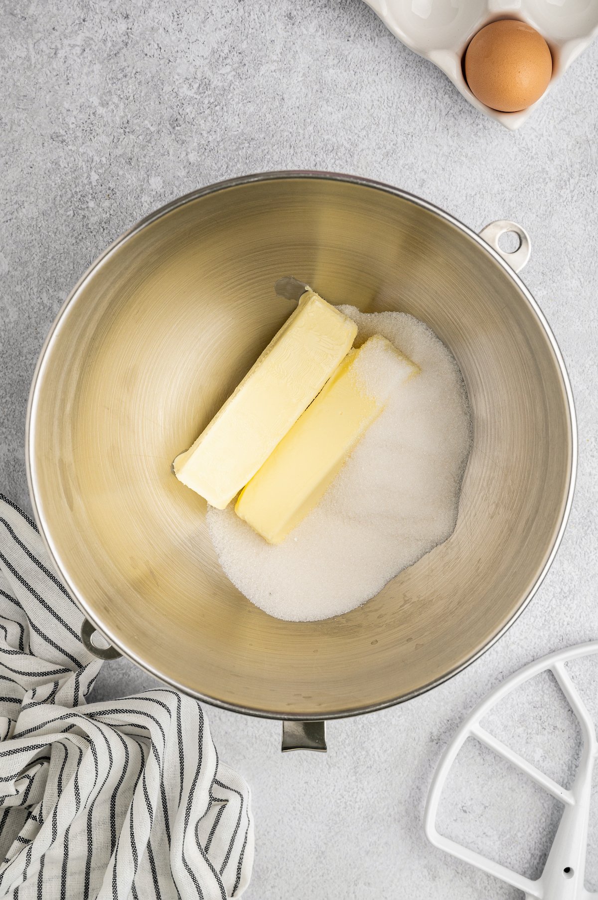 Sugar and butter in the bowl of a stand mixer. An egg in an egg carton is visible in the corner of the shot.