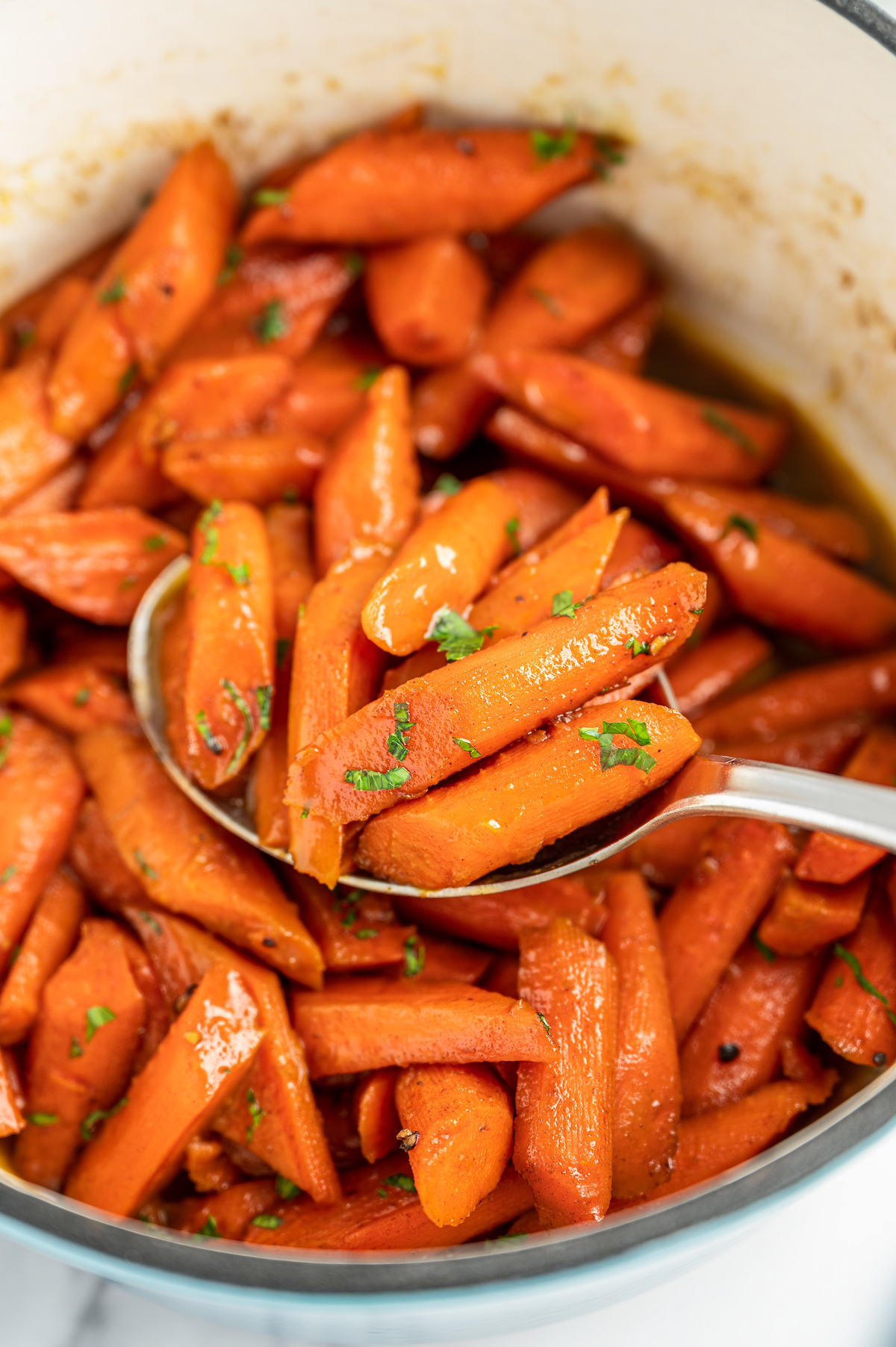 A serving spoon filled with candied carrots resting in a pot of carrots topped with fresh herbs.